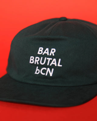 A dark green flat-brim five panel cap with white embroidered text reading 'BAR BRUTAL bCN' on the front, set against a solid red background.
