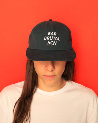A person wearing a dark green five panel cap with white embroidered text reading 'BAR BRUTAL bCN' against a solid red background.