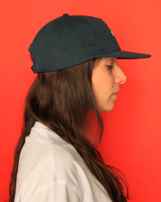 A side-profile portrait of a woman with long dark brown hair wearing a dark green five panel cap and a white shirt, set against a solid bright red background.