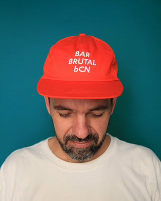 A man wearing a bright red baseball cap with white embroidered text reading 'BAR BRUTAL bcn' and a white t-shirt, set against a solid teal background.