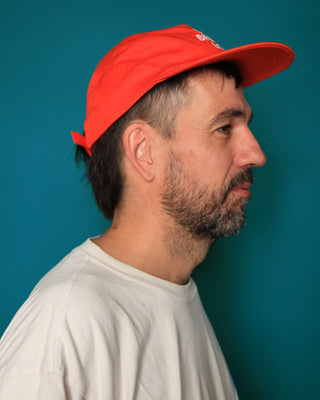 A man in profile wearing a bright red baseball cap and a light-colored t-shirt against a solid teal background.
