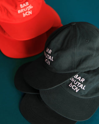 Two baseball caps on a teal background, one black in the foreground and one red partially visible behind it. Both caps have white embroidered text reading 'BAR BRUTAL BCN'.