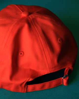 A close-up of a bright red baseball cap viewed from the back, showing the crown, button details, and adjustable black fabric strap with a metal slider, set against a dark teal background.