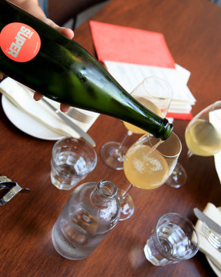 Person pouring white wine from a green bottle labeled 'Super" into a glass on a wooden table.