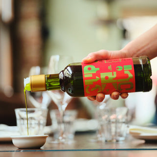 Person pouring a liquid from a bottle with a red label into a small bowl on a dining table.